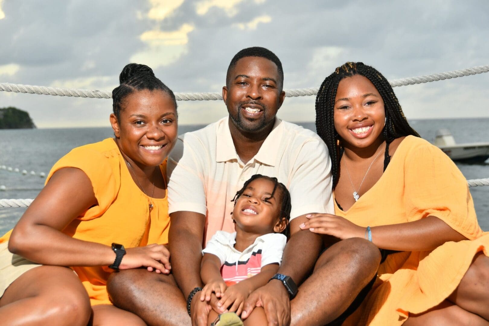 A family of four smiling and playing interactively for a photograph outdoors, dressed in coordinating colors by the water.